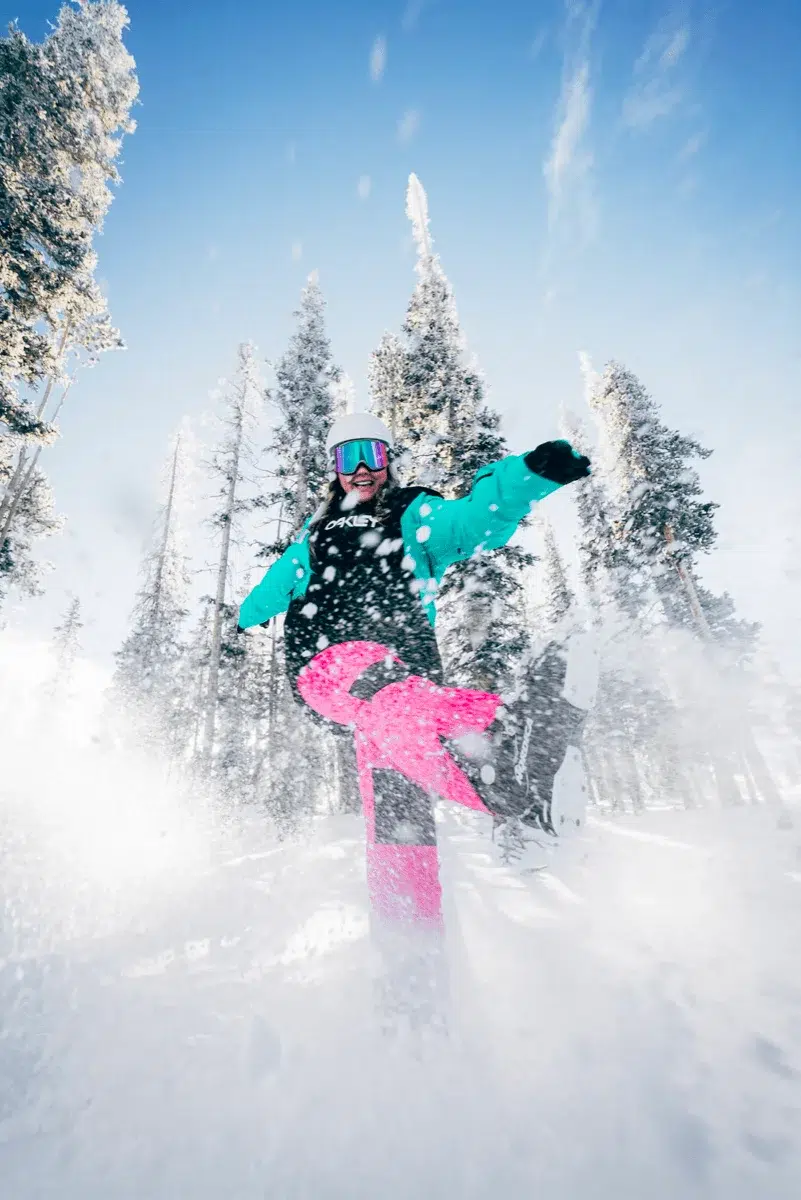 Een vrouw in ski-outfit, met roze skibroek, trapt losse sneeuw in de richting van de camera. Ze bevindt zich in een winters landschap waar skireizen voor bedrijven doorgaan, op een skivakantie met skipas.