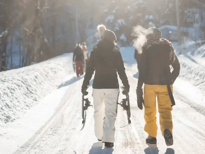 2 vrouwen wandelen in ski-outfit door de sneeuw. Ze hebben een snowboard in de hand. Mogelijks praten ze over het belang van verzekeringen op skireis.