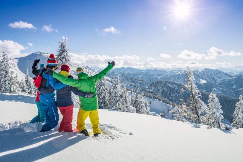 Een groep van vrienden, collega's of familieleden staat bovenaan een skipiste te poseren. Ze staan met hun rug naar de camera, kijken uit over het besneeuwde berglandschap en steken hun armen in de lucht.