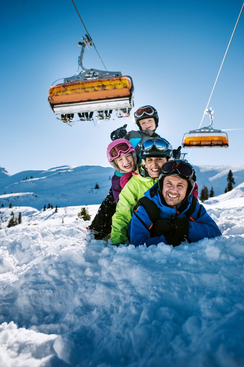 Een gezin van vier poseert in de sneeuw, op zonnige skivakantie met de familie. Ze liggen net onder de gele skilift.