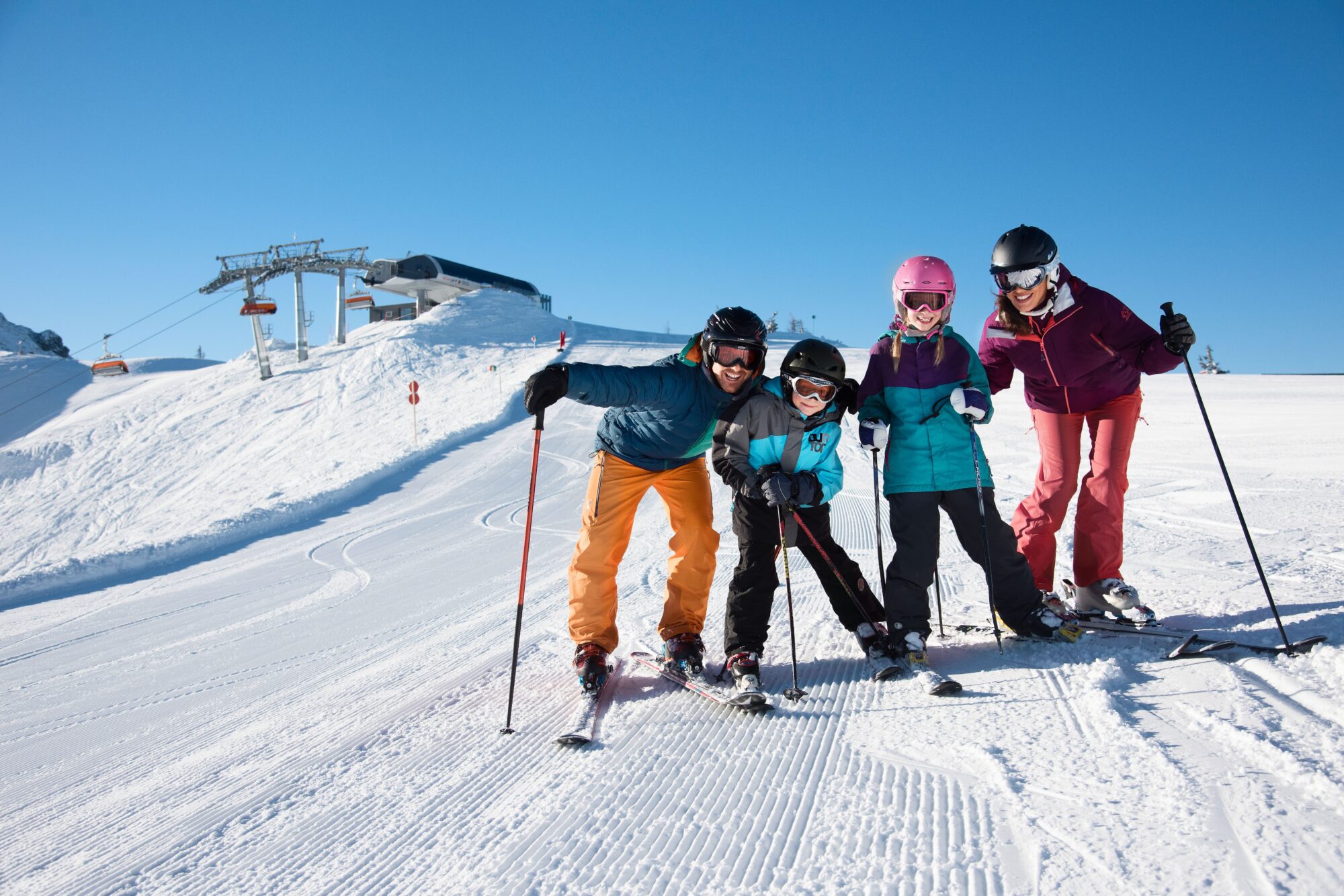Vier kinderen poseren voor de foto op een skipiste, op een heldere dag in de bergen. Ze hebben alle vier ski's aan. In de achtergrond zie je de skilift bovenkomen.
