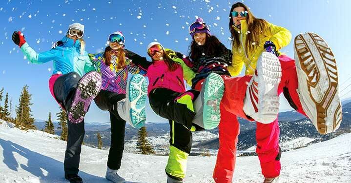 Een groep meisjes in ski-outfit in de sneeuw. Ze tonen de onderkant van hun sneeuwschoenen en lachen enthousiast. Ze zijn op een skireis voor jongeren met SnowCompanion