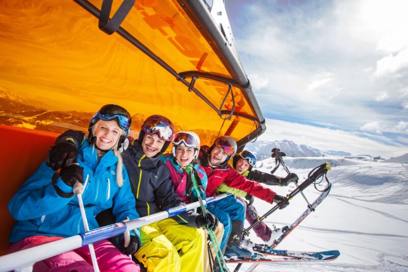 Een groep van vrienden die deelnemen aan een van de ski groepsreizen van SnowCompanion. Ze dragen allemaal een helm. Ze zitten in een skilift en poseren voor de foto.